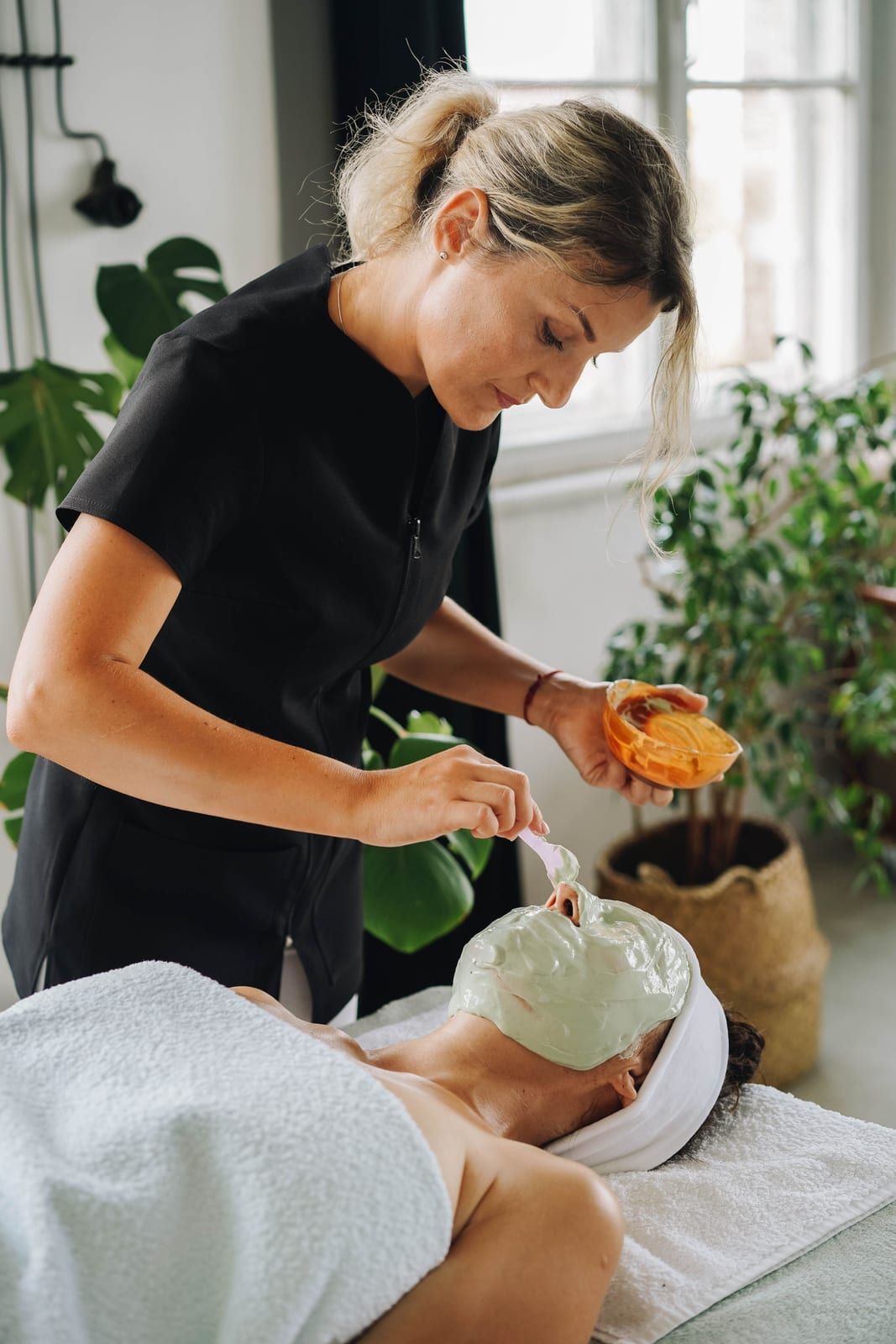 An aesthetician in black brushing a green clay mask onto a guest’s face in a bright room with plants