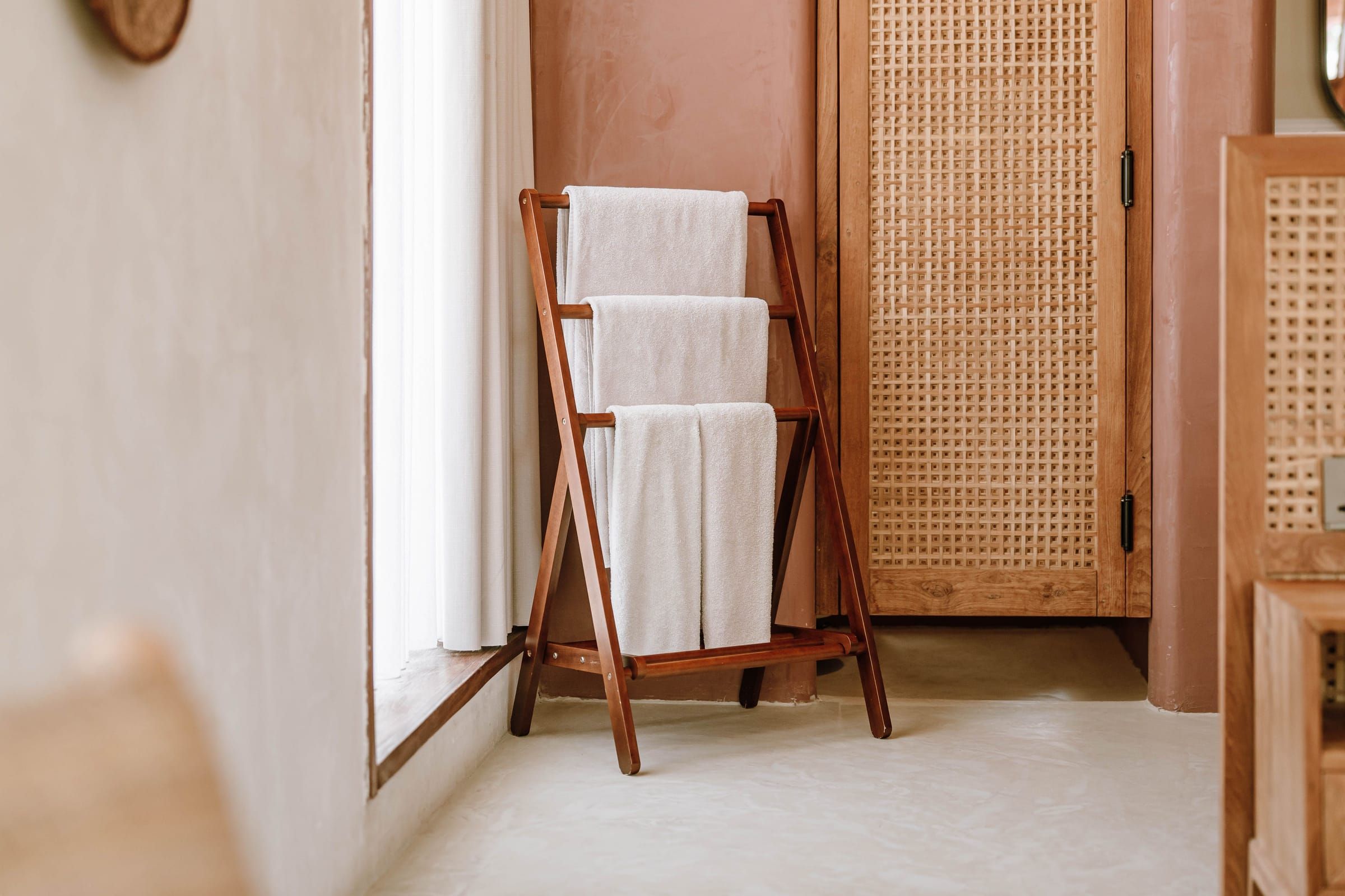 A wooden ladder rack hung with white towels beside a tall bright window, plaster walls and a woven cane door