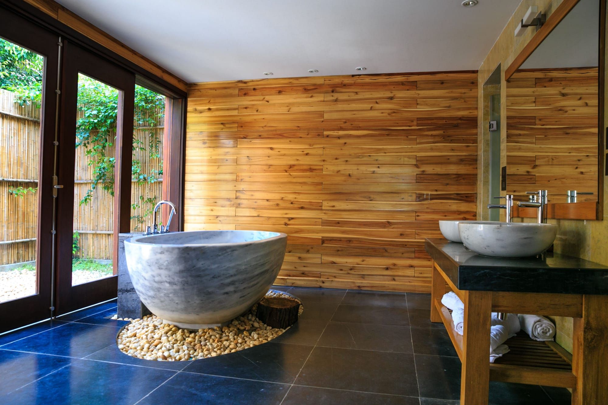 A round gray stone bathtub on a bed of pebbles in a timber-walled room, glass doors open to a garden